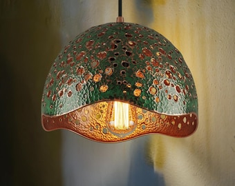 Może przedstawiać: Unikalna lampa wisząca z teksturowanym, zielonym i miedzianym kloszem. Klosz ma szereg małych otworów i falisty dolny brzeg. Żarówka jest widoczna wewnątrz, a lampa wisi na czarnym kablu.