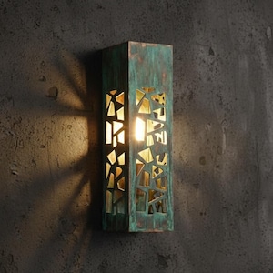 Peut inclure: Applique murale rectangulaire avec une finition en vert-de-gris. Le luminaire présente un motif géométrique avec des formes découpées qui laissent passer la lumière. Le luminaire est monté sur un mur gris foncé.