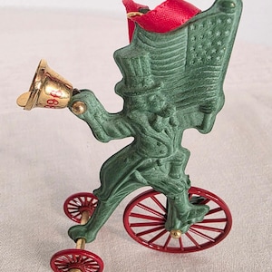 May include: A green Uncle Sam figure riding a penny-farthing bicycle with red wheels, holding an American flag. A gold-colored bell with the number 968 is attached to the figure's hand. A red ribbon loop is at the top for hanging as an ornament.