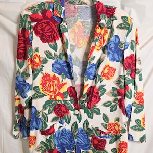 May include: A floral print blazer featuring a white background with large red, yellow, and blue rose patterns and green leaves. The blazer has a single-button closure and two front pockets. The long-sleeved jacket is hanging on a white hanger.