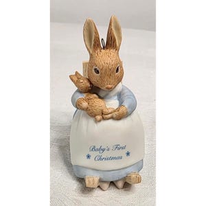 May include: A ceramic rabbit figurine wearing a light blue shirt and a white apron. The rabbit holds a smaller brown rabbit in its arms. The apron features the blue text Baby's First Christmas with small blue flower accents on each side.