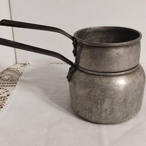May include: Vintage aluminum double boiler with two black handles. The pot has a cylindrical top section and a rounded bottom section. The handles are attached to the sides of the pot. The pot is sitting on a white surface.