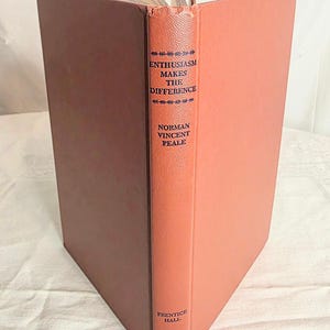 May include: An open book with a salmon-colored cover and spine. The spine features the title "Enthusiasm Makes the Difference" by Norman Vincent Peale. The book's pages are visible at the top, and the cover is a darker shade of brown.