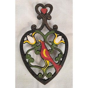 May include: A heart-shaped cast iron trivet featuring a dark textured finish with a central red bird perched among green vines and two white flowers with yellow accents. The top of the trivet includes a decorative scrollwork handle with a small red heart.