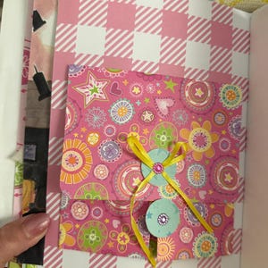 May include: A pink and yellow patterned envelope with a yellow ribbon and a blue and white button with a rhinestone. The envelope is decorated with stars, hearts, and flowers.