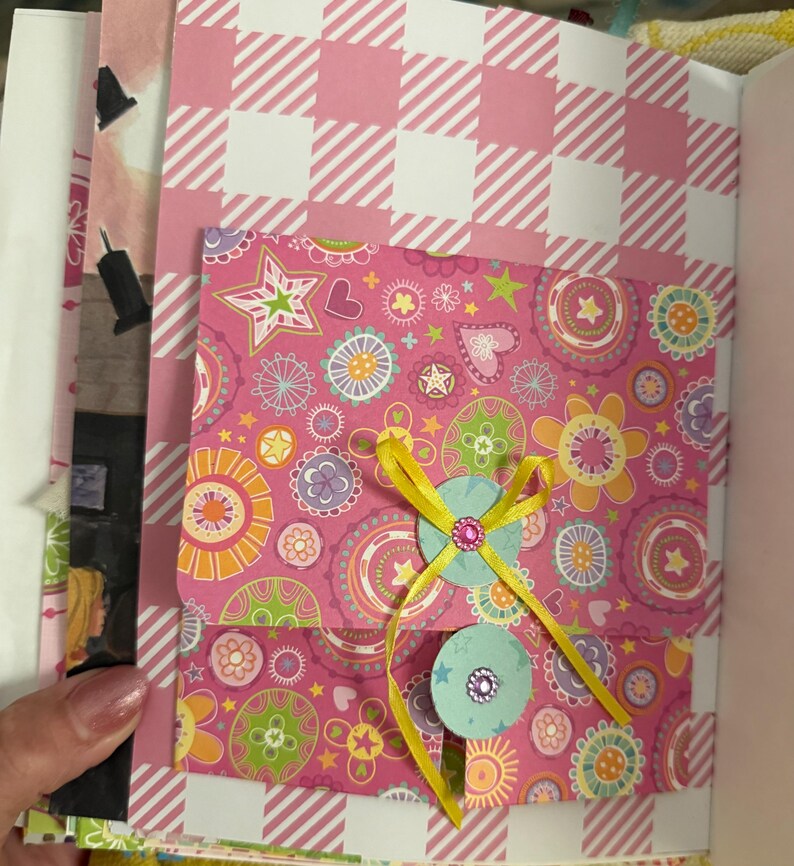 May include: A pink and yellow patterned envelope with a yellow ribbon and a blue and white button with a rhinestone. The envelope is decorated with stars, hearts, and flowers.