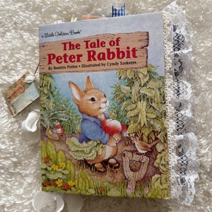 May include: A Little Golden Book edition of "The Tale of Peter Rabbit" by Beatrix Potter, illustrated by Cyndy Szekeres. The cover features a colorful illustration of Peter Rabbit holding a red tomato in a garden setting.