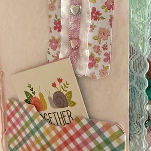 May include: A pink and white floral patterned ribbon with a scalloped edge and pink glitter ribbon is attached to a pink and white checkered pocket with the word "together" on a card inside. The pocket is attached to a pink journal with a green lace trim.