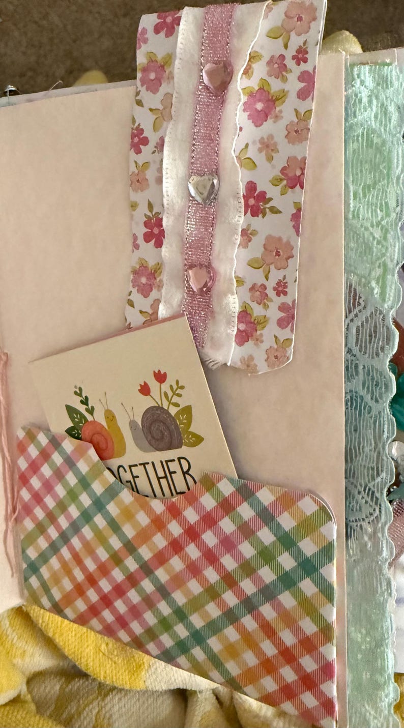 May include: A pink and white floral patterned ribbon with a scalloped edge and pink glitter ribbon is attached to a pink and white checkered pocket with the word "together" on a card inside. The pocket is attached to a pink journal with a green lace trim.