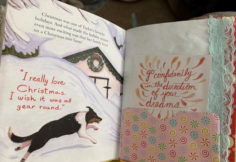 May include: An open book with a page featuring a dog running through the snow towards a house decorated for Christmas. The text on the page reads "I really love Christmas. I wish it was all year round." The opposite page has a pink background with a white card that reads "go confidently in the direction of your dreams".