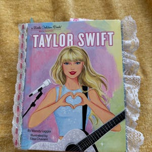 May include: A Little Golden Book biography of Taylor Swift. The cover features a colorful illustration of Taylor Swift with a microphone and guitar, making a heart shape with her hands. The book title is "Taylor Swift" and the subtitle is "Biography".