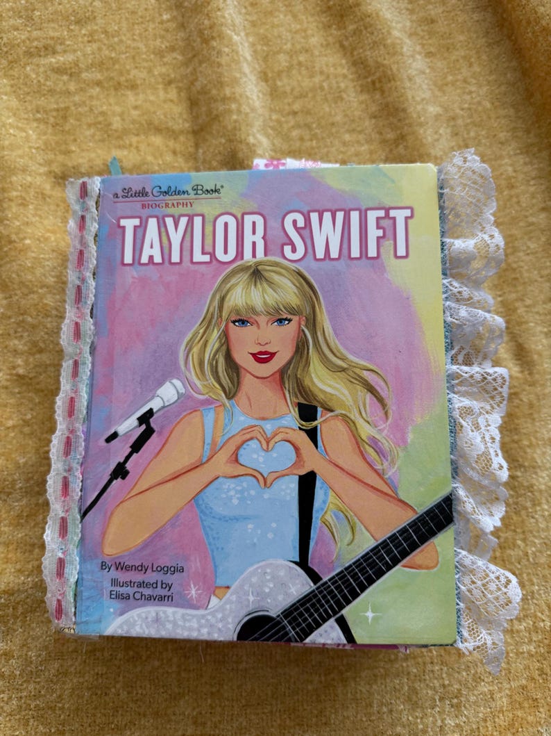 May include: A Little Golden Book biography of Taylor Swift. The cover features a colorful illustration of Taylor Swift with a microphone and guitar, making a heart shape with her hands. The book title is "Taylor Swift" and the subtitle is "Biography".
