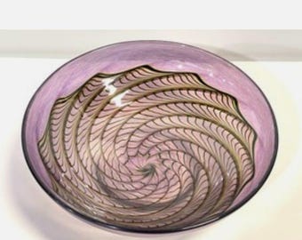 Può includere: Una ciotola in vetro circolare e poco profonda con una tonalità lavanda traslucida. La superficie interna presenta un motivo a spirale scuro e complesso che si irradia dal centro verso il bordo esterno, creando un design dettagliato a ragnatela.