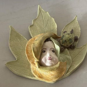 Op de afbeelding: Een decoratieve wandplaat met een bleek, gebeeldhouwd gezicht dat tevoorschijn komt uit een gekruld, goudbruin herfstblad. Het gezicht is omringd door grotere, lichtgroene, getextureerde bladeren, met een kleine, gedetailleerde bruine schildpad aan de rechterkant.