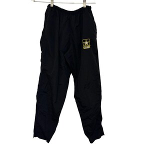 Puede incluir: Pantalón deportivo negro con cintura elástica, colgado de una percha metálica. La pierna derecha presenta un logotipo del Ejército de EE. UU. dorado. El pantalón está hecho de un material ligero, posiblemente nailon.