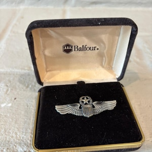Puede incluir: Pin de insignia militar plateado con alas, una estrella y un escudo, presentado en una caja de terciopelo negro abierta con ribete dorado. La caja muestra el logotipo de Balfour.
