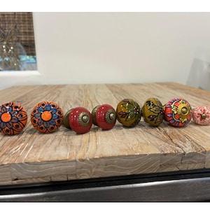 May include: A collection of decorative ceramic cabinet knobs in various colors and patterns. The knobs include floral and geometric designs in orange, blue, red, green, and yellow. Each knob has a metal base and is displayed on a wooden surface.
