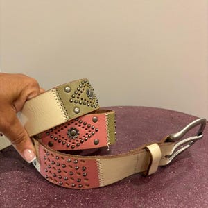 Op de afbeelding: Een veelkleurige leren riem met een zilveren gesp en decoratieve studs. De riem is gemaakt van drie verschillende gekleurde lederen panelen: crème, groen en roze. De riem heeft een zilveren gesp en is versierd met zilveren studs.