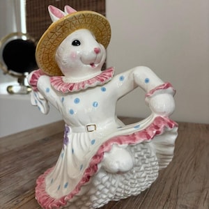 May include: A white ceramic bunny figurine wearing a straw hat and a white dress with blue polka dots and pink trim. The bunny is posed as if dancing, with one arm raised and the other holding up the dress.