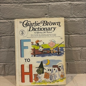 May include: Vintage "The Charlie Brown Dictionary" Volume 3 book. The cover features Snoopy and Charlie Brown fishing, and Halloween-themed illustrations. The book's title and author are printed in black, with the letters "F to H" in blue and red.