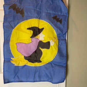 May include: A blue Halloween flag featuring a witch flying on a broomstick in front of a yellow moon. Two black bats are flying in the sky.