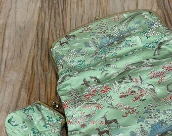 Puede incluir: Un conjunto de bolso de mano y monedero verde menta. La tela presenta un diseño de paisaje detallado con ciervos, árboles y pájaros en tonos verdes, rojos y azules. El bolso de mano tiene un cierre dorado.