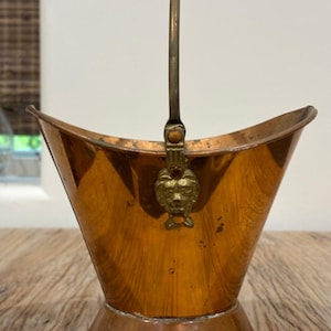 Könnte beinhalten: Ein kupferfarbener Eimer mit Griff und einem dekorativen Löwenkopf-Emblem. Der Eimer hat einen ausgestellten oberen Rand und einen runden Sockel. Der Griff ist aus dunklerem Metall. Der Eimer steht auf einer Holzoberfläche.