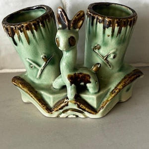 Peut inclure: Un pot de fleurs vintage en céramique avec une glaçure vert clair. Le pot présente deux ouvertures cylindriques et une figurine de lapin centrale. Des accents bruns mettent en valeur les bords et les détails du lapin. Le pot a un design unique et fantaisiste.