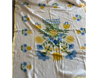 Op de afbeelding: Een wit deken met een gele rand en een bloemenpatroon in tinten blauw, geel en groen. Het ontwerp bevat een vaas met bloemen en bladeren.