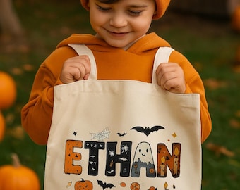 May include: A beige Halloween tote bag with the name "Ethan" in orange and black letters. The bag features Halloween-themed graphics, including a ghost, bats, and pumpkins. The person holding the bag is wearing an orange beanie and hoodie.
