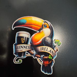 Könnte beinhalten: Ein farbenfroher Aufkleber mit einem Tukan, der auf einem Ast sitzt und ein Pint Guinness hält. Die Wörter "Guinness" und "Ireland" sind auf Bannern zu sehen. Ein Kleeblatt ist ebenfalls im Design enthalten. Der Tukan hat einen großen orange-gelben Schnabel.