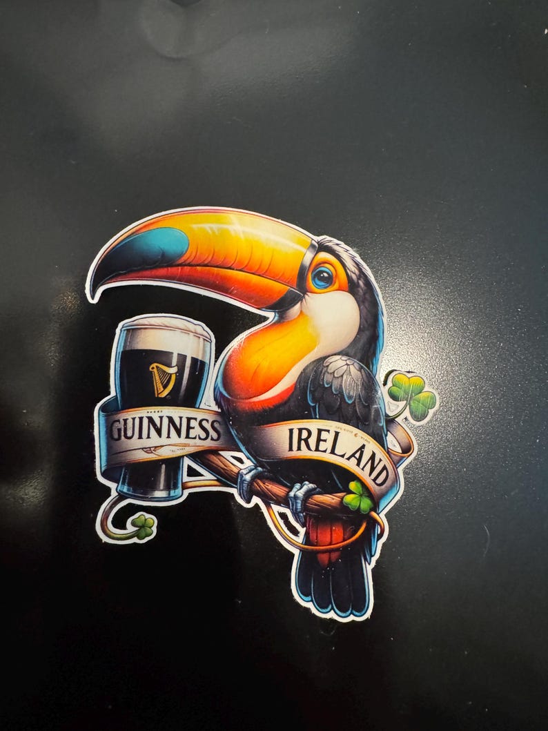 May include: A colorful sticker featuring a toucan perched on a branch, holding a pint of Guinness. The words "Guinness" and "Ireland" are displayed on banners. A shamrock is also included in the design. The toucan has a large orange and yellow beak.