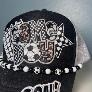 May include: Black trucker hat with the words "GAME DAY" in a soccer-themed design. The hat features a beaded band with soccer ball accents and the word "GOAL!" on the brim. The hat has a black glitter brim and a white mesh back.