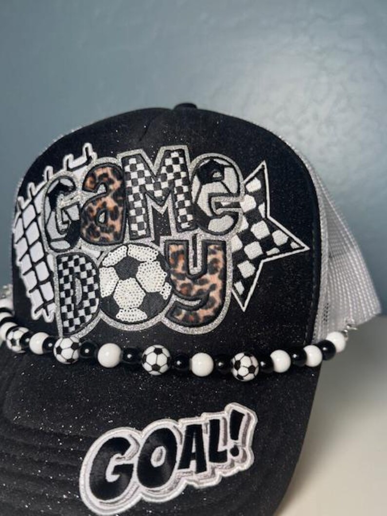 May include: Black trucker hat with the words "GAME DAY" in a soccer-themed design. The hat features a beaded band with soccer ball accents and the word "GOAL!" on the brim. The hat has a black glitter brim and a white mesh back.