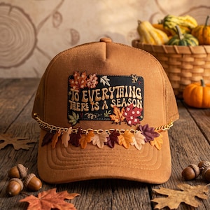 May include: A tan trucker hat featuring a rectangular patch with embroidered autumn leaves and the text "TO EVERYTHING THERE IS A SEASON". A gold-toned chain is draped across the front, adorned with small fabric leaves in shades of orange, red, and brown.