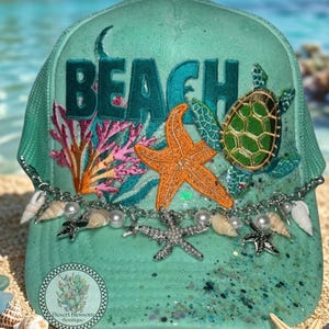May include: A teal trucker hat with the word "BEACH" embroidered on the front. The hat is decorated with a starfish, sea turtle, coral, and seashells. A chain with pearls and starfish charms hangs from the brim.