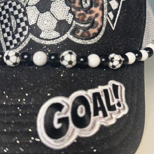 May include: Black glitter baseball cap with a soccer ball design and the word "GOAL!" in white. The cap features a beaded band with soccer ball beads and a checkered flag design.