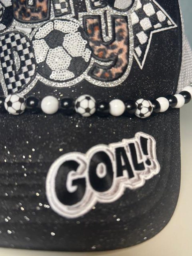 May include: Black glitter baseball cap with a soccer ball design and the word "GOAL!" in white. The cap features a beaded band with soccer ball beads and a checkered flag design.