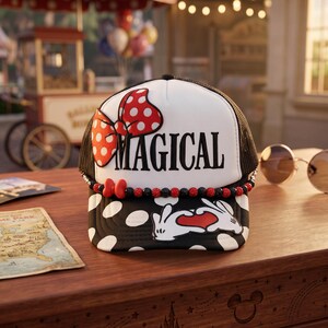 May include: A white and black trucker hat featuring a red and white polka dot bow, the word MAGICAL in black lettering, a beaded trim with red and black spheres, and a graphic of white cartoon gloves forming a heart shape on the black brim.