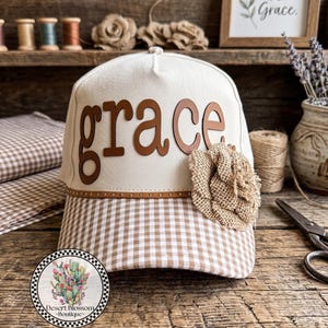 May include: A cream-colored baseball cap with the word "grace" in brown letters. The brim features a brown and white gingham pattern. A decorative burlap flower is attached to the side. The cap is displayed on a wooden surface.