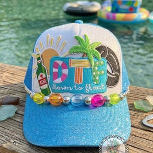 May include: A blue and white trucker hat with a glittery blue brim. The hat features a design with a sun, palm tree, and the words "DT down to float". A beer bottle graphic is also included.