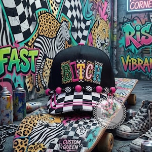 May include: A black baseball cap with a checkered brim and pink piping, featuring a beaded trim and the word BITCH in colorful, patterned letters. The cap sits on a skateboard with a leopard and zebra print design, surrounded by spray paint cans.