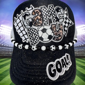 May include: Black baseball cap with the words "GAME DAY" and "GOAL!" in white. The letters "GAME DAY" have a soccer ball and checkered flag design. A beaded band with soccer ball accents encircles the cap.