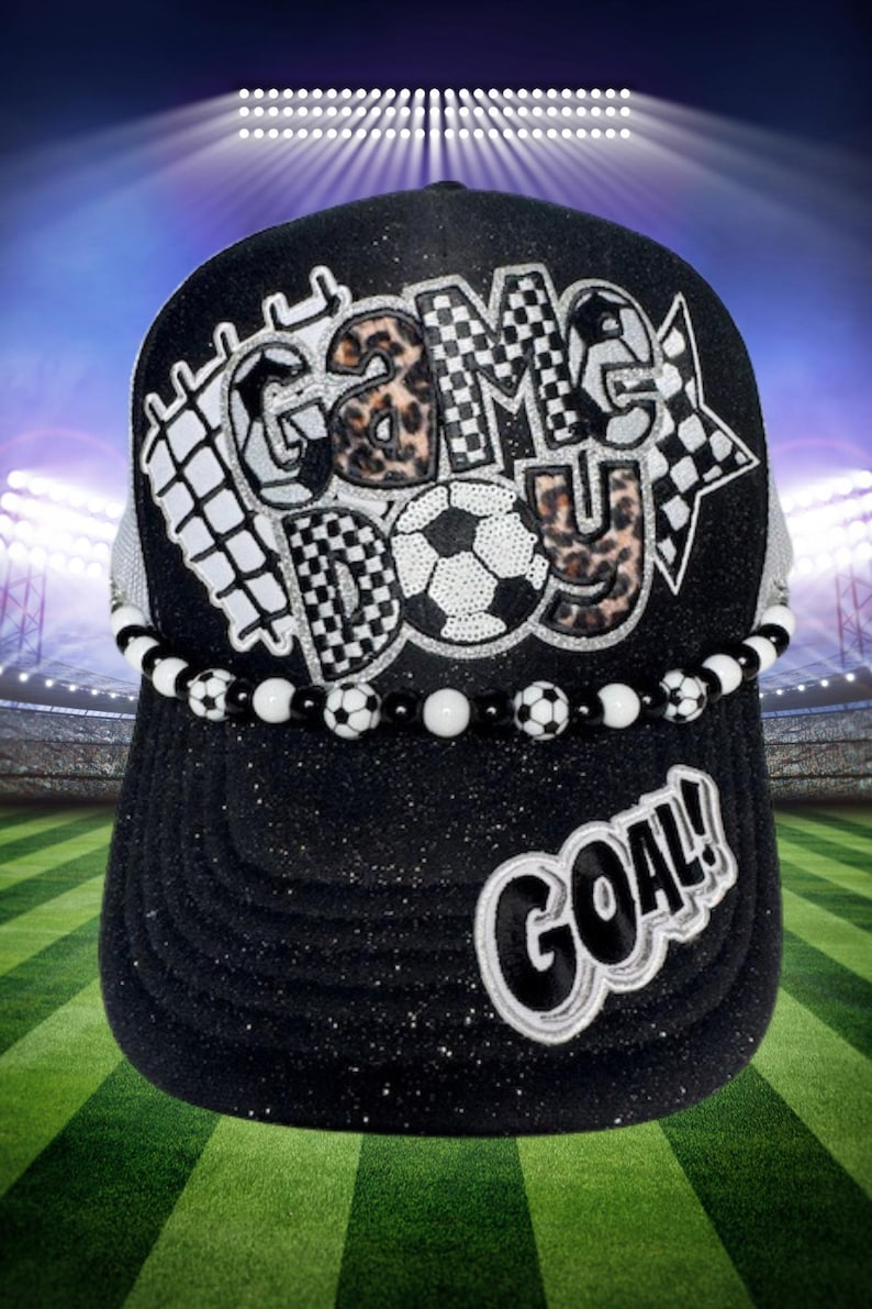 May include: Black baseball cap with the words "GAME DAY" and "GOAL!" in white. The letters "GAME DAY" have a soccer ball and checkered flag design. A beaded band with soccer ball accents encircles the cap.