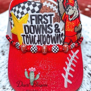May include: Red baseball cap with a football theme. Features the text "First Downs & Touchdowns" and a cartoon wolf in a KC jersey. Includes a star, a checkered flag, and a cactus logo.