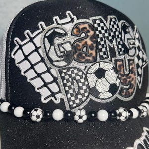 May include: Black glitter baseball cap with the words "GAME DAY" in a mix of patterns, including leopard print and soccer balls. The brim is decorated with a beaded chain of soccer balls and white and black beads. The cap has a white mesh back.