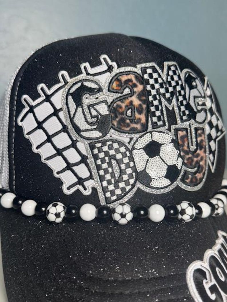 May include: Black glitter baseball cap with the words "GAME DAY" in a mix of patterns, including leopard print and soccer balls. The brim is decorated with a beaded chain of soccer balls and white and black beads. The cap has a white mesh back.