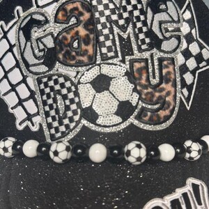 May include: Black baseball cap with the words "GAME DAY" in a mix of leopard print, checkered, and silver glitter designs. A soccer ball is in the center. A beaded band with soccer ball beads and white beads is at the base. The word "GOAL!" is at the bottom.
