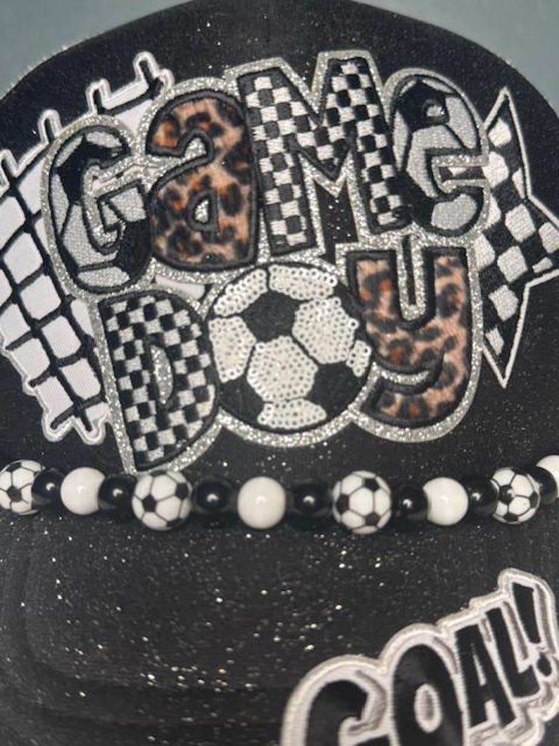 May include: Black baseball cap with the words "GAME DAY" in a mix of leopard print, checkered, and silver glitter designs. A soccer ball is in the center. A beaded band with soccer ball beads and white beads is at the base. The word "GOAL!" is at the bottom.