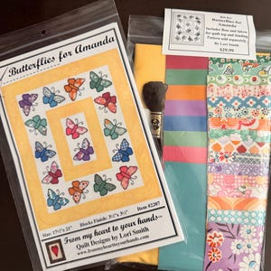May include: A craft kit titled Butterflies for Amanda by Lori Smith, featuring a pattern sheet with a yellow-bordered quilt design showing colorful butterflies and a separate plastic bag containing assorted fabric strips and embroidery floss.